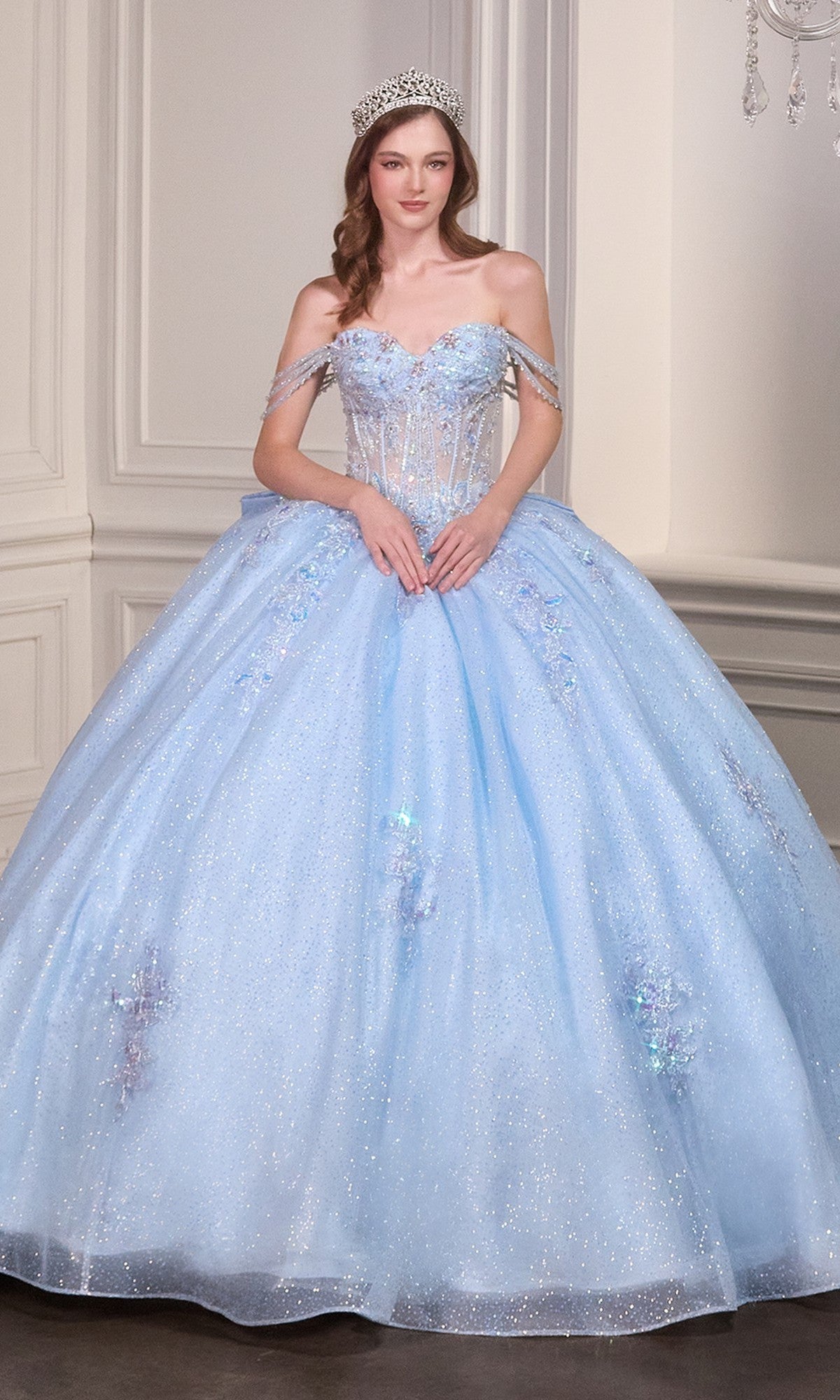 A woman in the Quinceanera Dress 15735 by Ladivine, a sparkling light blue ball gown with an off-the-shoulder neckline and tiara, stands in an ornate room with white paneled walls.